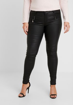 Persoon die zwarte skinny neplederen broek draagt met ritssluitingdetails, gecombineerd met zwarte open teen sandalen met hoge hak en een lichtroze top met lange mouwen.