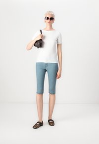 Bela kratka majica s kratkimi rokavi, svetlo modri jeans capri hlače, črna torba, rjave čevlje s tigrastim vzorcem in veliki sončna očala. Enobarvna ozadje.