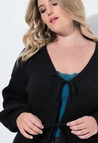 Svart stickad kofta med långa ärmar, öppen framsida och knytning. Buren över en teal topp med spetsdetaljer. Texturerad tygdetalj synlig.
