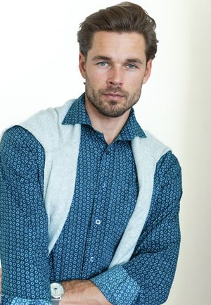 Junger Mann mit blau gemustertem Hemd und hellgrauem Pullover, der über den Schultern drapiert ist, schaut selbstbewusst in die Kamera, die Arme verschränkt.