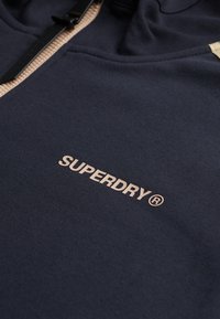 Marineblauwe capuchontrui van zacht stof, voorzien van een volledige ritssluiting en een beige logoopdruk met de tekst "SUPERDRY" aan de voorkant.