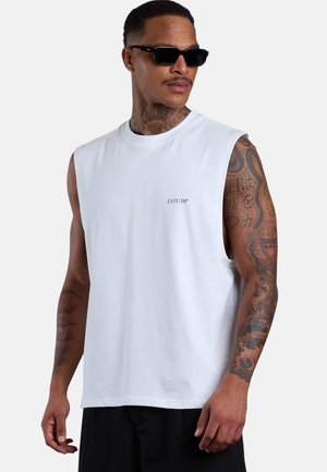 Man wearing black sunglasses and a white sleeveless shirt with tattoos on both arms against a plain background.