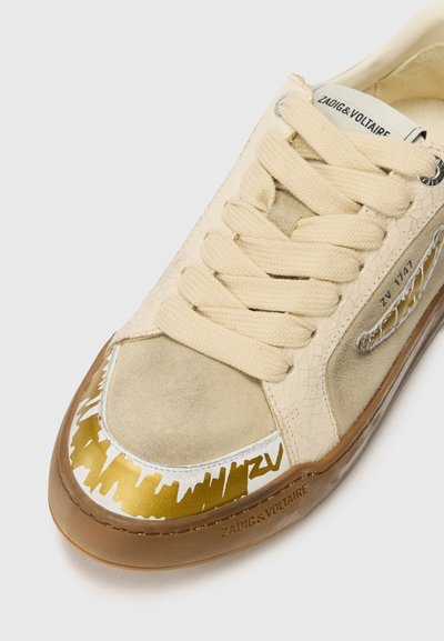 Beżowy sneaker z teksturowaną cholewką ze zamszu i popękanej skóry, kremowe sznurowadła oraz złoty abstrakcyjny wzór na nosku. Gumowa podeszwa.