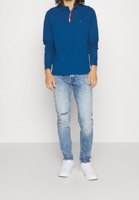 Modrá polokošile s dlouhým rukávem s červenobílým lemováním límce, doplněná světle modrými džínami s efektem opotřebení a bílými teniskami.