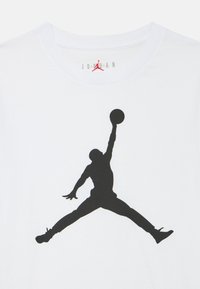 Biała bawełniana koszulka z dużym czarnym sylwetowym obrazem skaczącej postaci trzymającej piłkę do koszykówki. Nad dekoltem znajduje się małe czerwone logo.
