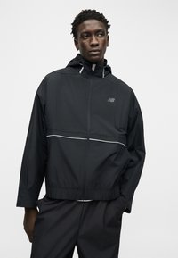 Άνδρας φορώντας μαύρο μπουφάν με κουκούλα και φερμουάρ, με λευκές ραφές και λογότυπο New Balance, με τα χέρια στις τσέπες, κοιτάζοντας ελαφρώς προς τα δεξιά πάνω σε απλό φόντο.