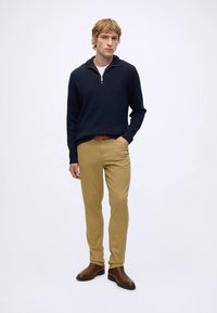 Uomo biondo in piedi con maglione blu navy con zip, canottiera bianca, pantaloni beige con cintura marrone e stivali di pelle marroni su sfondo bianco semplice.