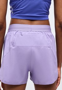 Vue arrière d'une personne portant un short de sport lavande avec une poche zippée et un haut de sport violet.