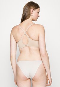 Frau trägt beige Kreuzrücken-BH und helle Unterwäsche, von hinten vor einem schlichten weißen Hintergrund gezeigt.