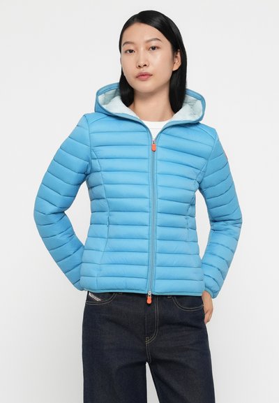 Veste matelassée bleue avec un design en quilt horizontal, fermeture éclair complète et capuche, dotée d'un tirette de fermeture éclair orange et d'une doublure bleu clair.