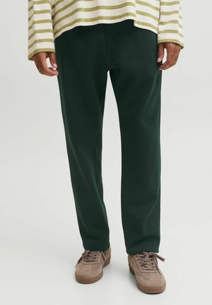 Personne portant un pantalon vert foncé, des baskets beiges et une chemise blanche à manches longues avec des rayures horizontales vert clair.
