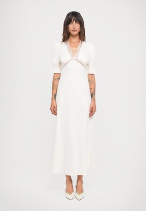 Witte jurk met pofmouwen, v-hals en kantdetails. Aansluitende lijfje vloeit over in een lange rok. Gecombineerd met witte puntige schoenen.