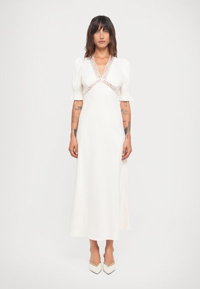 Weißes Kleid mit Puffärmeln, V-Ausschnitt und Spitzenverzierungen. Enganliegendes Oberteil fließt in einen langen Rock über. Kombiniert mit weißen spitzen Schuhen.