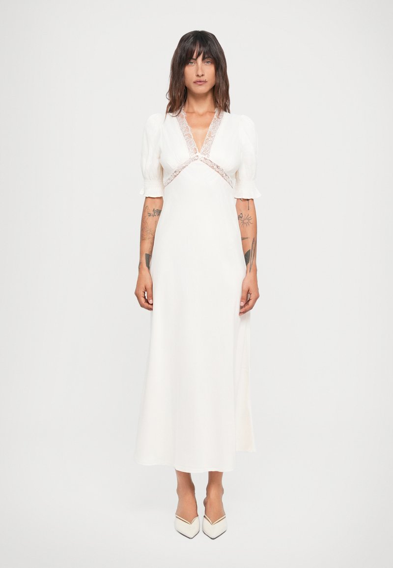 Witte jurk met pofmouwen, v-hals en kantdetails. Aansluitende lijfje vloeit over in een lange rok. Gecombineerd met witte puntige schoenen.