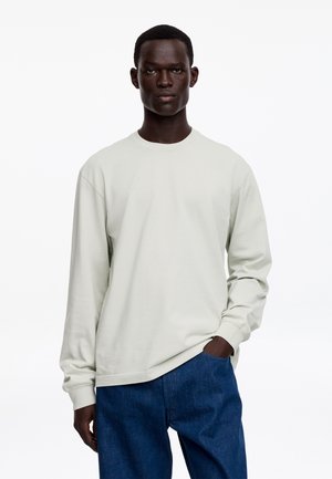 Młody mężczyzna o ciemnej karnacji, ubrany w białą koszulę z długim rękawem i niebieskie dżinsy, stojący z jedną ręką w kieszeni na jednolitym tle.