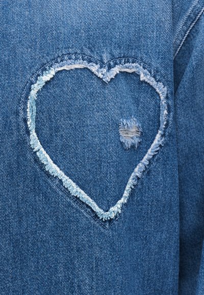 Écusson en forme de cœur effiloché avec un petit trou usé au centre sur un tissu en denim bleu.