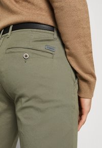 Gros plan d'une personne portant un pantalon vert olive avec une poche arrière à bouton et un pull marron à manches longues rentré, montrant une ceinture noire.