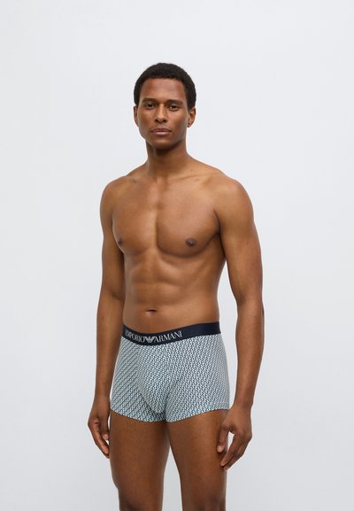 Μπόξερ στενά με μπλε ζώνη μέσης που φέρει το λογότυπο "Emporio Armani". Το ύφασμα είναι διακοσμημένο με μικρά μαύρα τρίγωνα σε ανοιχτό φόντο.