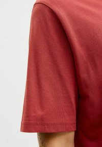 Rød bomulds T-shirt med korte ærmer, der har en glat tekstur, rund hals og en pænt syet kant. Nærbillede af skulderen og ærmet.