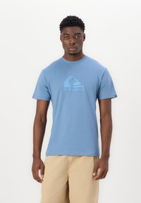 Blå kortærmet bomulds t-shirt med et lyseblåt grafisk logo på brystet. Rund halsudskæring og afslappet pasform. Ensfarvet baggrund.