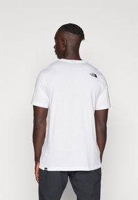 Hvit t-skjorte med korte ermer laget av bomull, med en svart logo på ryggen. Rund hals og løs passform. Kombinert med mørke shorts.