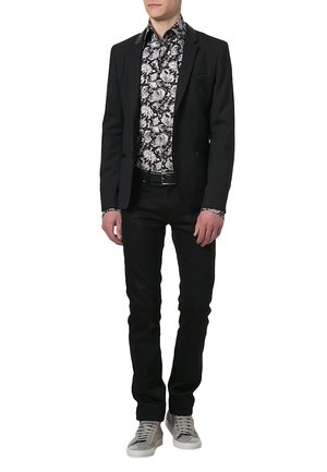 Homme portant un blazer noir, une chemise noire à fleurs blanches, un pantalon noir, une ceinture noire et des baskets grises, debout devant un fond blanc.