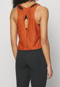 Žena v bezramínkovém oranžovém texturovaném crop topu s průstřihovými zády, pod kterým má černou sportovní podprsenku, a černých legínách.