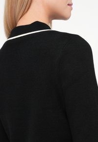 Femme aux cheveux blonds portant un pull noir avec une bordure blanche le long du col, vue de dos et de profil.