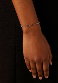 Zilveren kettingarmband met verlengde schakel en een decoratieve middelste schakel. Draagt op een hand met een natuurlijke huidskleur.