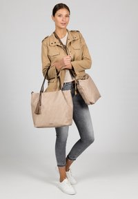 Beige tote bag with a textured finish and tassel accent, paired with a smaller matching bag. Model wears a light brown jacket and gray jeans.