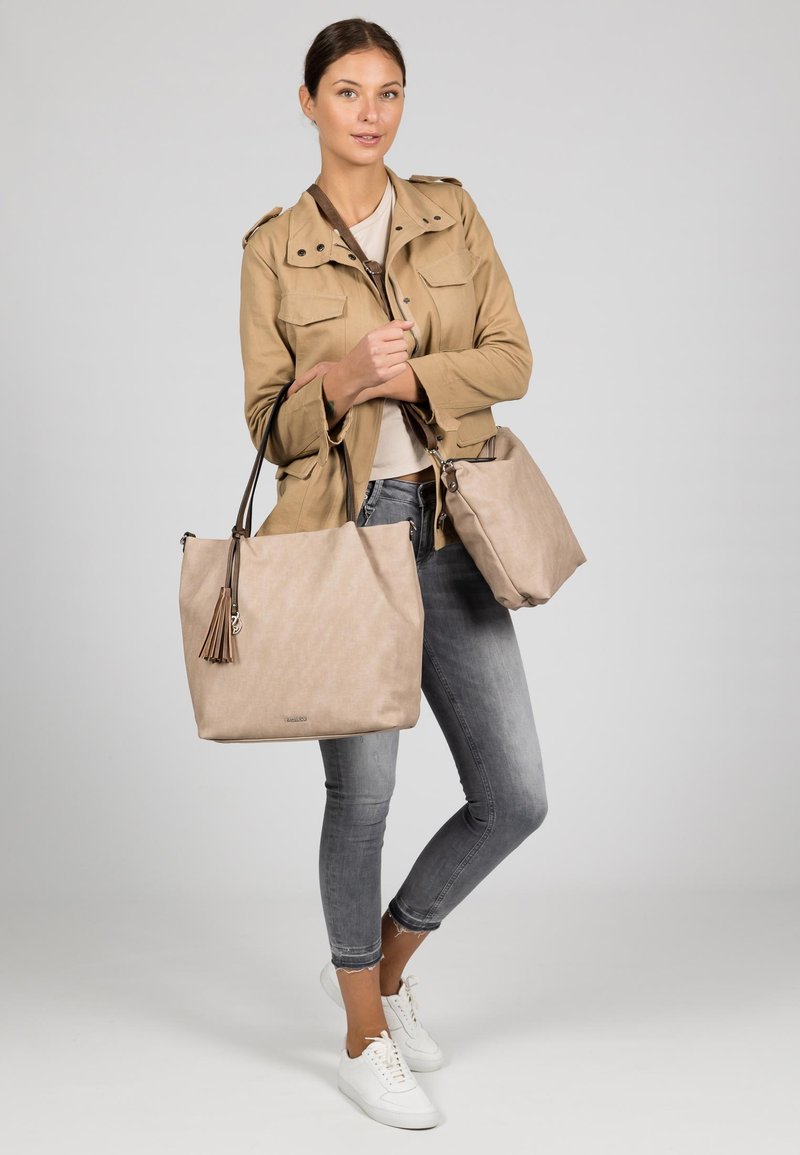 Beige tote bag with a textured finish and tassel accent, paired with a smaller matching bag. Model wears a light brown jacket and gray jeans.