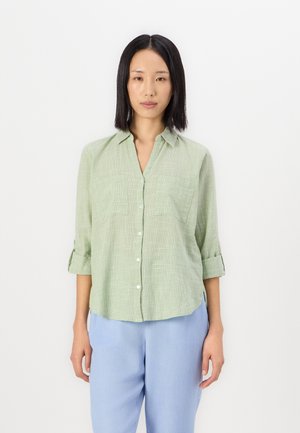 Chemise verte clair à manches roulées, avec deux poches poitrine et un tissu texturé. Associée à un pantalon bleu clair.