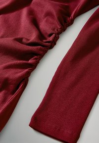 Bordeauxfarbenes geripptes Material mit gerafften Abschnitten und langen Ärmeln, das Textur und eine glatte Oberfläche zeigt. Das Design weist einen dezenten Stretch auf.