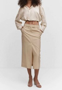 Blouse beige boutonnée associée à une jupe crayon beige longueur genou avec une fente à l'avant. Tissu à texture lisse et coupe décontractée.