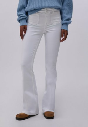 Persona che indossa pantaloni bianchi a zampa d'elefante a vita alta, maglione a coste azzurro chiaro e scarpe marroni con punta quadrata su sfondo neutro.