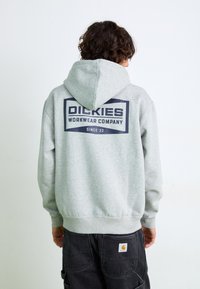 Hanorac gri cu un logo grafic mare de culoare bleumarin pe spate, având inscripțiile "Dickies Workwear Company" și "Din '22." Mâneci și tiv ribbate.