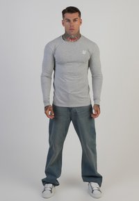 Hosszú ujjú szürke póló, kerek nyakkivágással és fehér logóval a mellkasán; világoskék farmernadrággal és fehér sportcipővel párosítva.