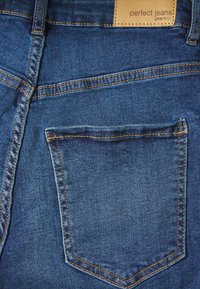 Džínové kalhoty v tmavě modré barvě s kontrastním zlatým prošíváním. Obsahují obdélníkovou zadní kapsu a značkový štítek našitý na pasu.