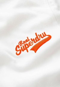 Valge kangas, millel on ringikujuline logo, mille peal on bold-kirjas oranž tekst "Real Superdry", millele lisab kaarekujuline allajoon.