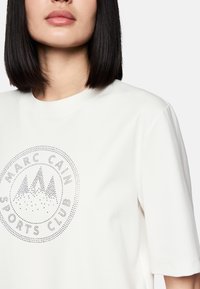 White short-sleeve t-shirt made of soft fabric featuring a circular silver dotted logo design with mountain graphics and text.