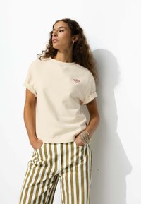 T-shirt en coton beige avec des manches retroussées et un petit logo rouge et bleu. Associé à un pantalon rayé vert et blanc.