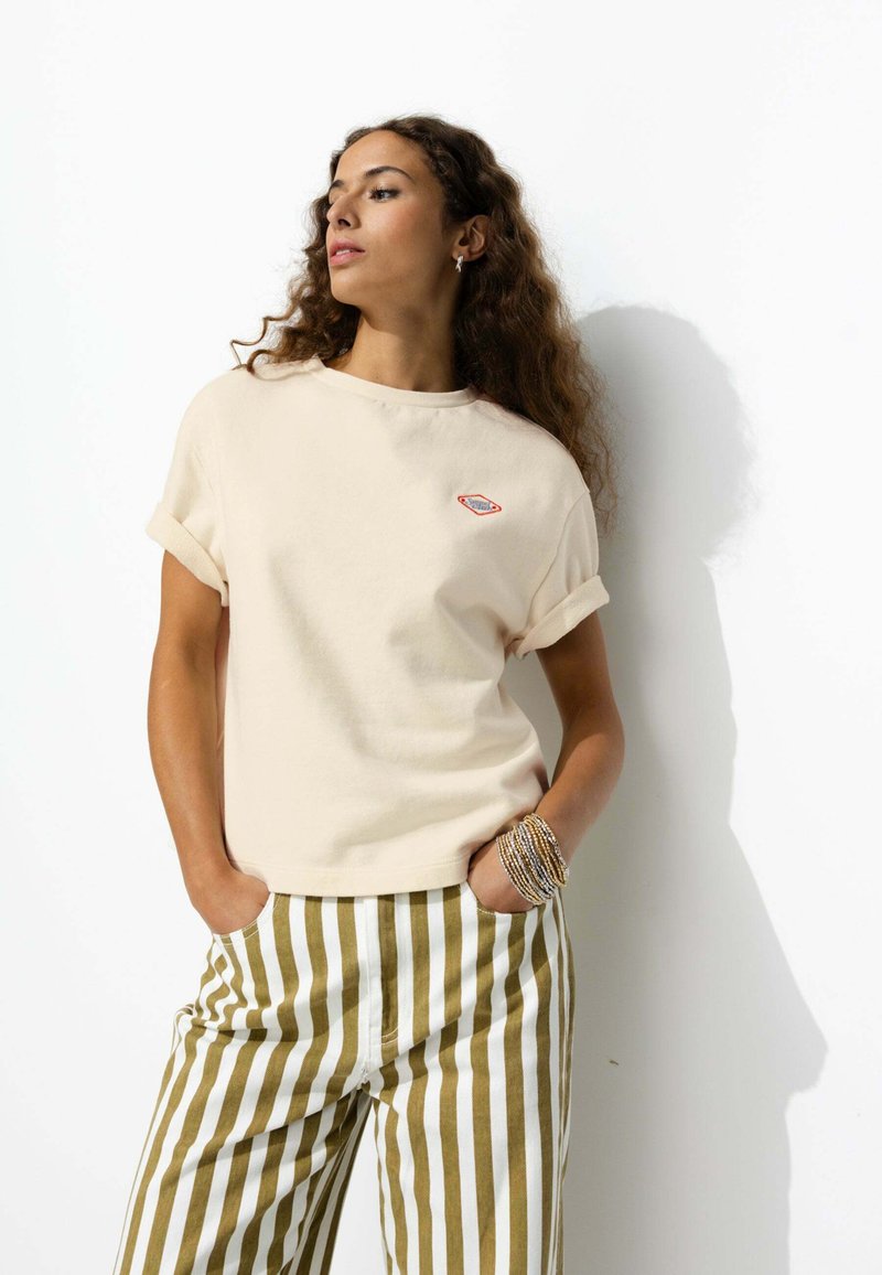 T-shirt en coton beige avec des manches retroussées et un petit logo rouge et bleu. Associé à un pantalon rayé vert et blanc.