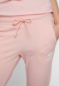 Ljusrosa sweatpants i mjukt tyg, med ett dragsko i midjan och en vit logotyp på nedre benet. Slät textur genom hela plagget.