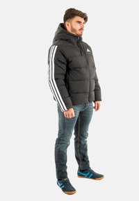 Chaqueta acolchada negra con capucha, que presenta un destacado diseño de tres rayas blancas en las mangas. Combinada con jeans azules y zapatillas negras.