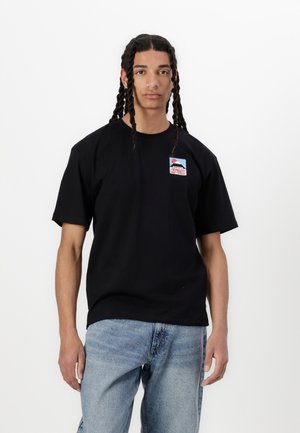 T-shirt nera a maniche corte con collo a giro, caratterizzata da un patch logo colorato sul lato sinistro del petto. Indossato con jeans in denim azzurri.