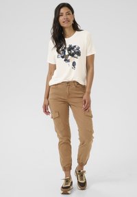 Helles beige T-Shirt mit navyblumigem Druck; beige Cargo-Hosen mit elastischen Bündchen; braune und cremefarbene Sneaker. Einfaches, lässiges Outfit.