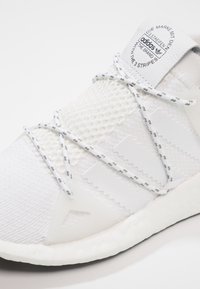 Valkoiset sneakerit, joissa on verkko- ja teksturoitu kangas, harmaat nauhat, kumiset yksityiskohdat ja logotagi kielen päällä. Kevyt muotoilu.