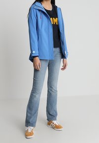 Persona con chaqueta azul, camiseta negra con letras amarillas, jeans de luz azul y zapatillas naranjas y blancas de pie contra un fondo liso.