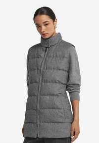 Kvinna som bär en grå vadderad ärmlös väst med hög krage och dragkedja, över en långärmad grå tröja, stående mot en vit bakgrund.