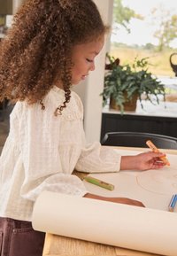 Child with curly hair drawing on a large cream paper sheet. Surrounding are colorful markers, soft lighting, and indoor greenery.